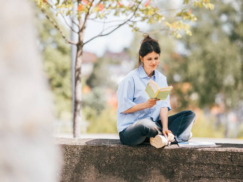 Eine Frau sitzt draußen auf einer Mauer und liest ein Buch.