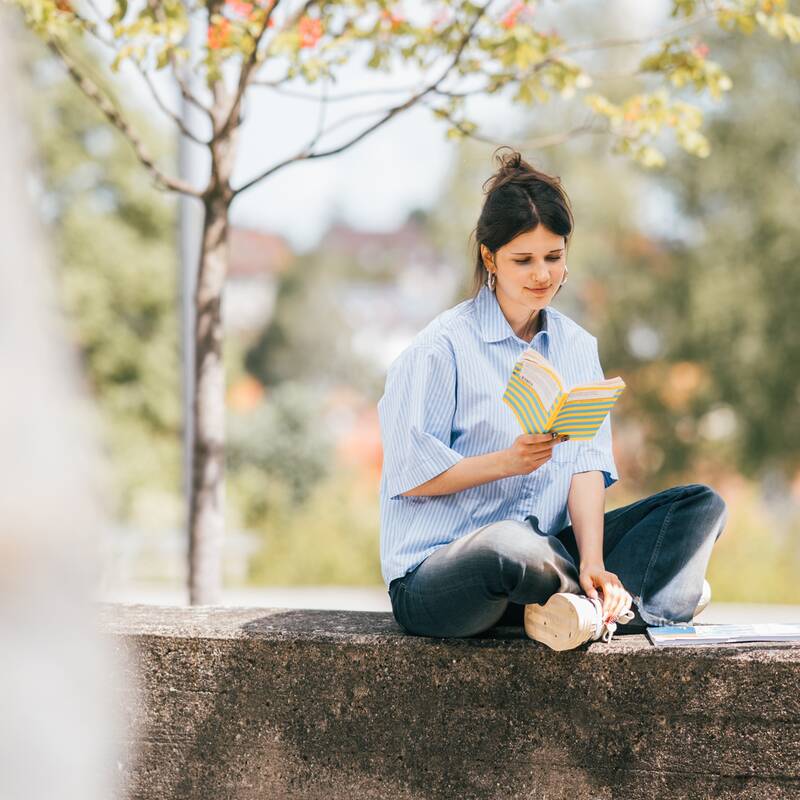 Eine Frau sitzt draußen auf einer Mauer und liest ein Buch.