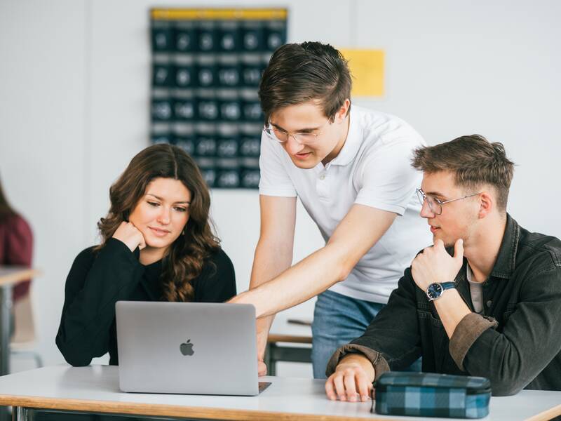 Ein Mann zeigt zwei Personen etwas auf einem Laptop in einem Klassenzimmer.