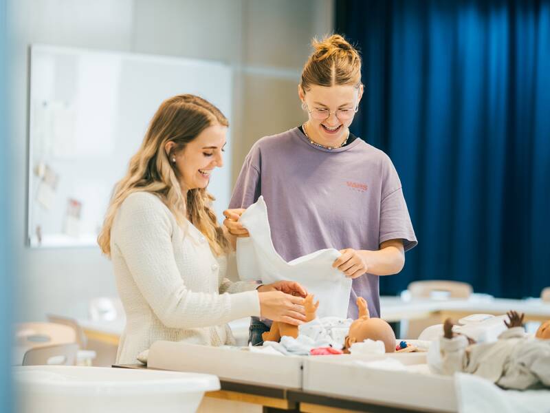 Zwei Frauen stehen an einem Tisch und üben Windelwechseln an einer Babypuppe.