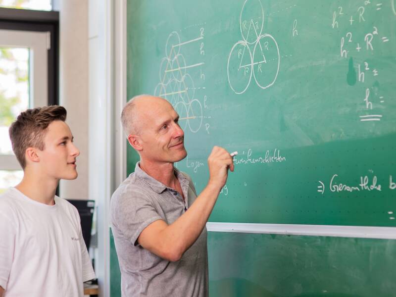 Ein Lehrer erklärt einem Schüler mathematische Formeln an einer grünen Tafel.
