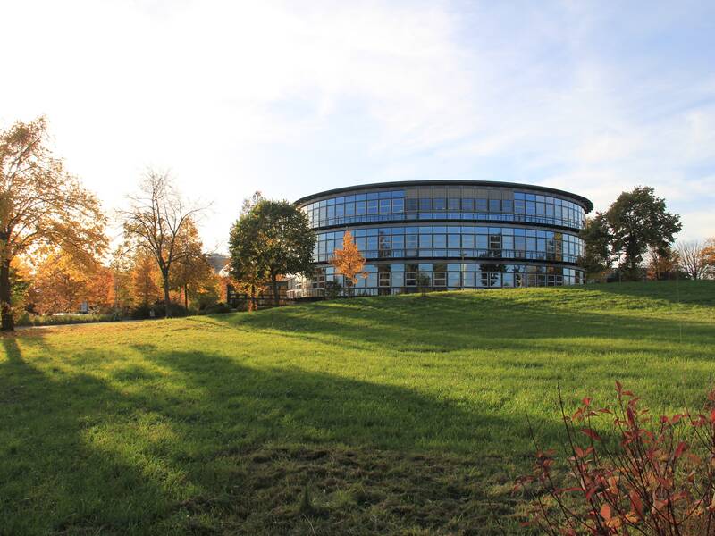 Ein modernes, rundes Gebäude mit vielen Fenstern steht auf einer grünen Wiese, umgeben von herbstlich gefärbten Bäumen.