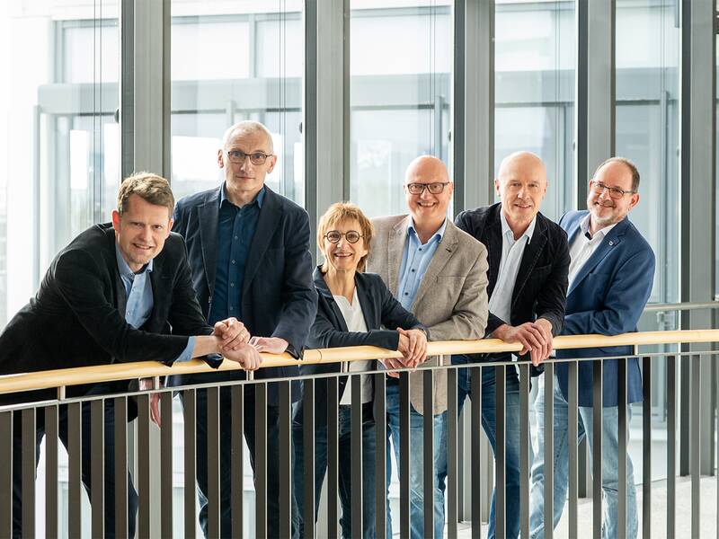 Sechs Personen, vier Männer und zwei Frauen, stehen lächelnd in einem modernen Büroflur und tragen Business- und Freizeitkleidung.