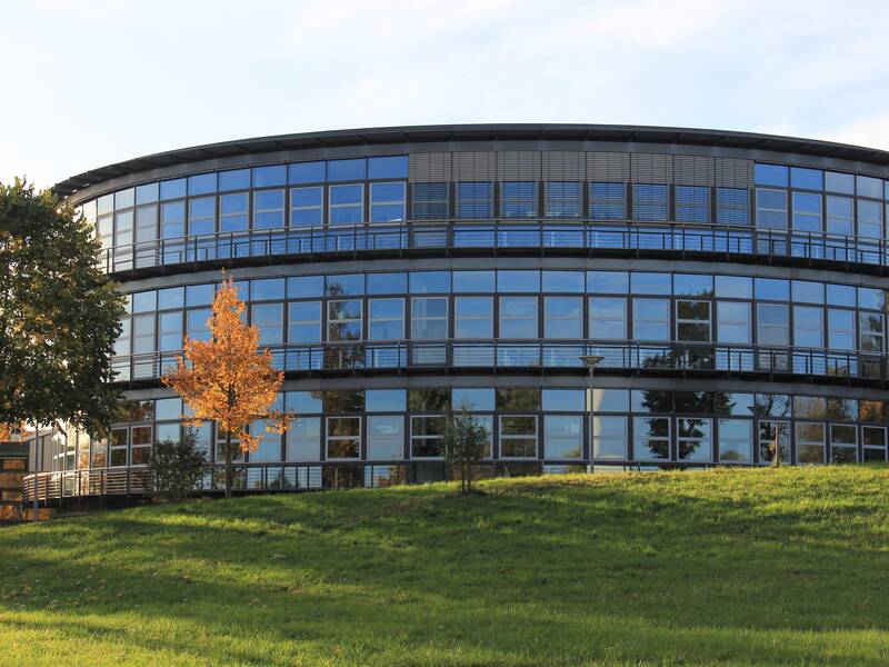 Ein modernes, rundes Gebäude mit vielen Fenstern steht auf einer grünen Wiese, umgeben von einigen Bäumen im Herbstlaub.