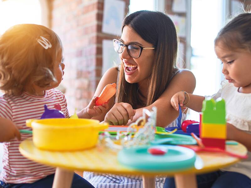 Eine Frau spielt freudig mit zwei kleinen Kindern an einem Tisch mit buntem Spielzeug.