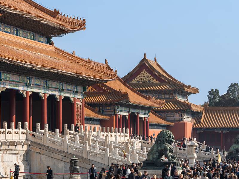 Das Bild zeigt einen Teil der Verbotenen Stadt in Peking mit traditionellen chinesischen Gebäuden und einer Menschenmenge im Vordergrund.