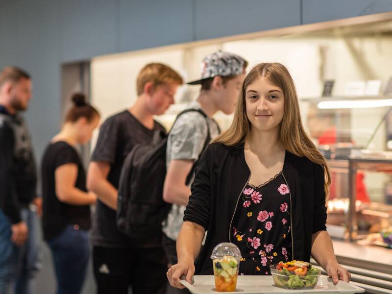 Eine junge Frau, die ein Tablett mit einem Salat und einem Obstbecher trägt, steht lächelnd in einer Cafeteria, während mehrere Menschen in der Schlange dahinter stehen.
