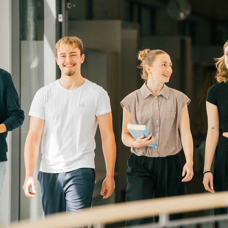 Vier junge Erwachsene gehen entspannt und lachend in einem modernen Gebäude mit Glastüren.