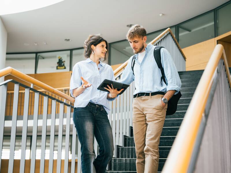 Zwei Personen gehen eine Treppe hinunter und unterhalten sich, während eine der beiden ein Tablet in der Hand hält.