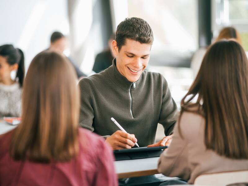 Ein lächelnder junger Mann sitzt an einem Tisch und schreibt mit einem Stift auf einem digitalen Gerät, während andere Personen im Hintergrund arbeiten oder sprechen.