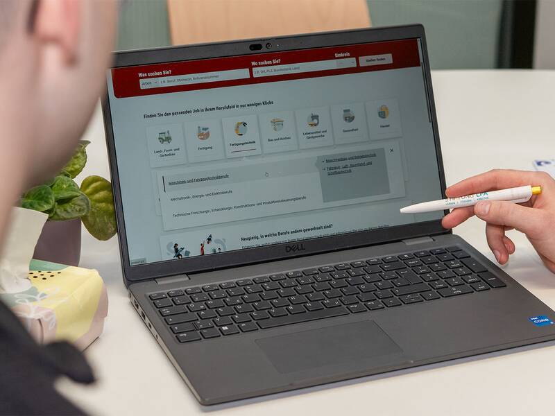 Eine Person zeigt mit einem Stift auf den Bildschirm eines Laptops, auf dem eine Webseite mit Suchoptionen angezeigt wird, während eine andere Person daneben sitzt.