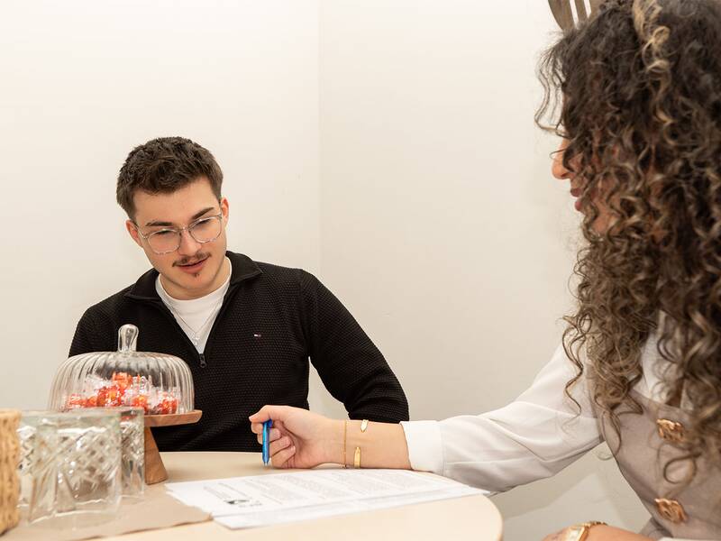 Ein Mann und eine Frau sitzen an einem Tisch mit Papierkram, bunten Stiften und einer Schale mit Bonbons, während sie miteinander sprechen.