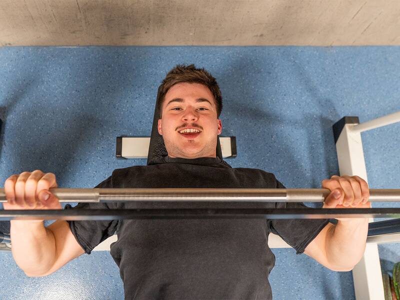 Eine Person liegt auf einer Bank in einem Fitnessstudio und hebt eine Langhantelstange.