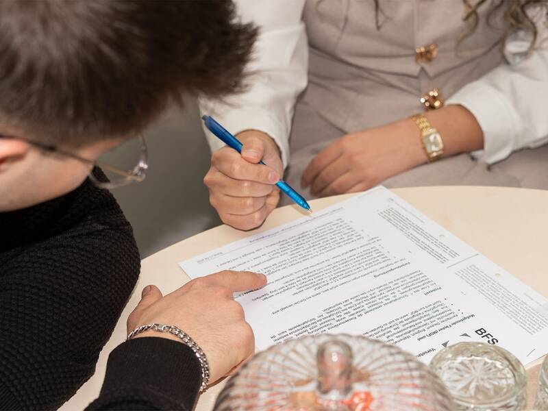 Zwei Personen sitzen an einem Tisch und studieren gemeinsam Dokumente, wobei eine Person etwas mit einem blauen Stift markiert.