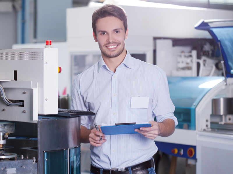 Ein lächelnder Mann steht in einer Fabrikumgebung, hält ein Klemmbrett und trägt ein weißes Hemd.