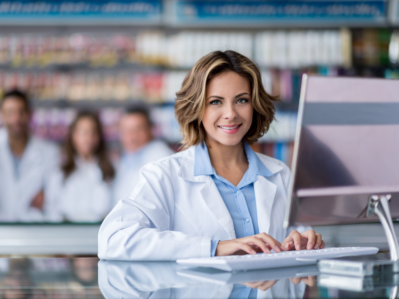 Eine Frau in einem weißen Kittel sitzt lächelnd an einem Computer in einer Apotheke, während mehrere Personen im Hintergrund zu sehen sind.