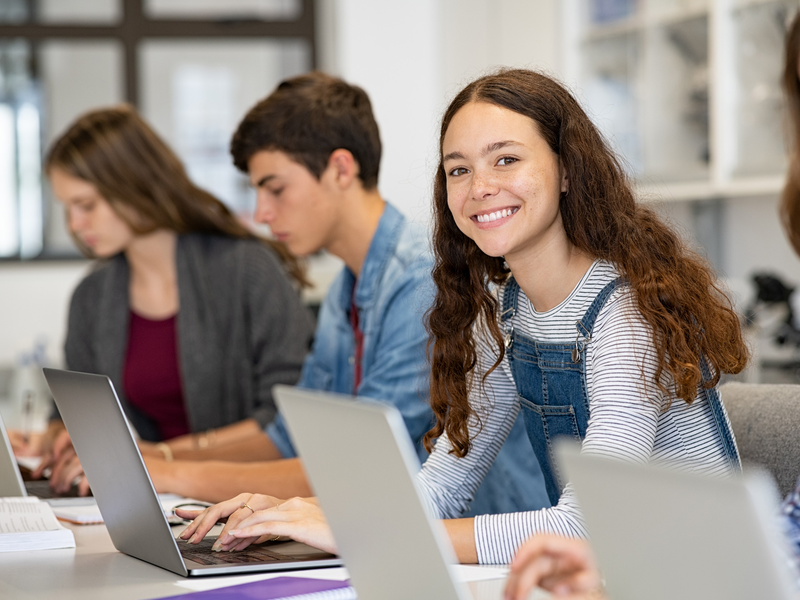 Eine Gruppe von Jugendlichen arbeitet in einem Klassenzimmer an Laptops, wobei eine Person in der Mitte lächelnd in die Kamera schaut.