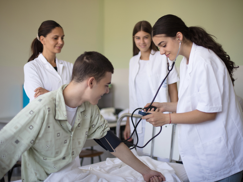 Eine junge Medizinstudentin misst den Blutdruck eines sitzenden Patienten, während zwei weitere Personen in weißen Kitteln zusehen.