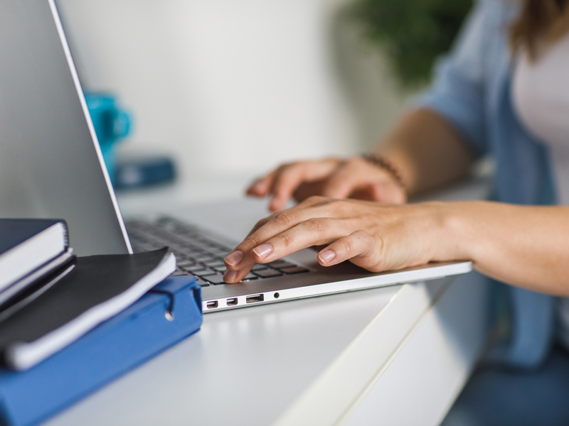 Eine Person tippt auf einer Laptop-Tastatur, die auf einem weißen Schreibtisch neben einem Stapel Bücher steht.