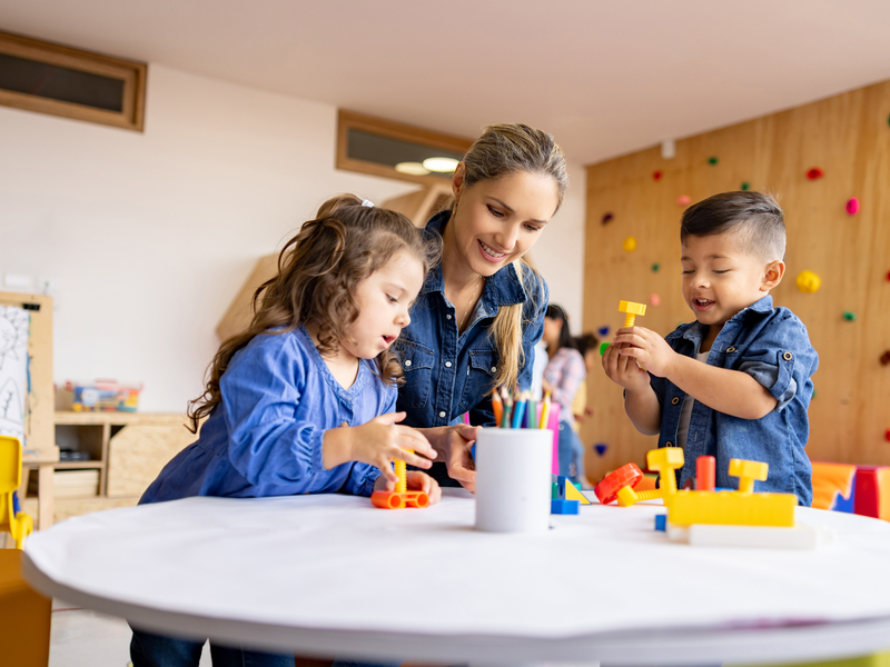 Eine Erzieherin sitzt mit zwei Kindern an einem Tisch in einer bunt dekorierten Umgebung und sie spielen zusammen mit Bausteinen.