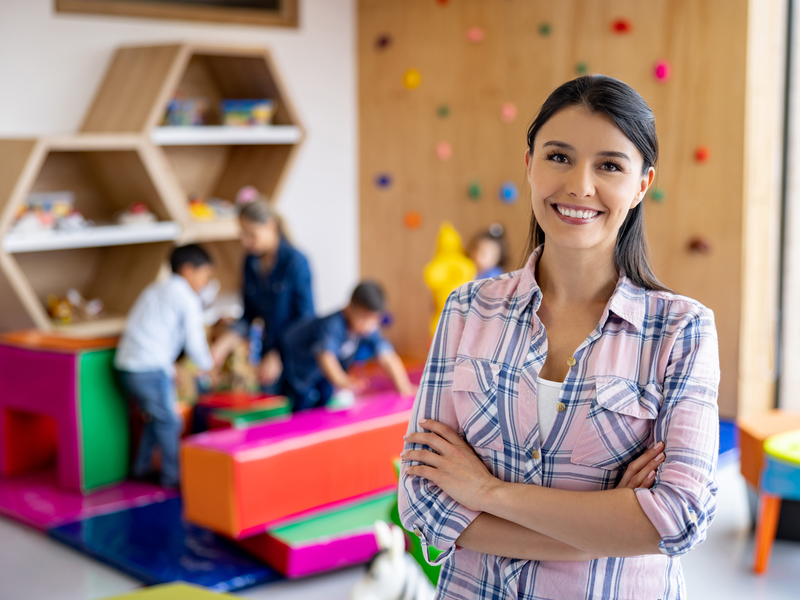 Eine lächelnde Frau steht mit verschränkten Armen im Vordergrund eines farbenfrohen Spielzimmers, während Kinder im Hintergrund spielen.