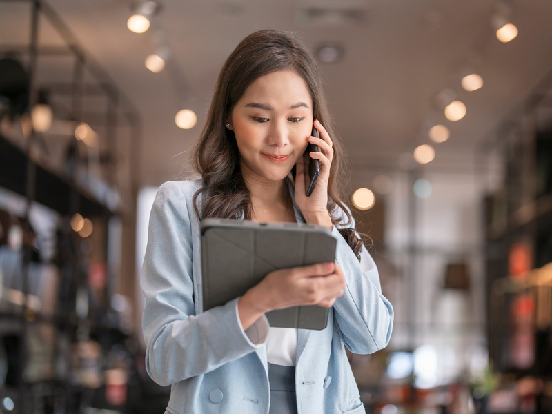 Eine Frau in heller Kleidung steht in einem Geschäft, spricht in ein Smartphone und hält ein Tablet.