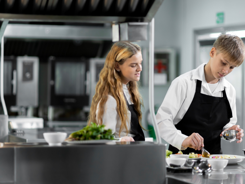 In einer professionellen Küche bereiten zwei Jugendliche in weißen Hemden und schwarzen Schürzen gemeinsam eine Mahlzeit zu.