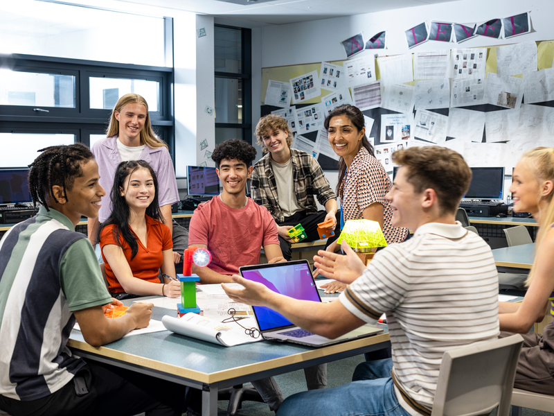 Eine Gruppe von jungen Erwachsenen sitzt und steht um einen Tisch in einem Klassenzimmer, schaut fröhlich und scheint an einem Projekt zu arbeiten.