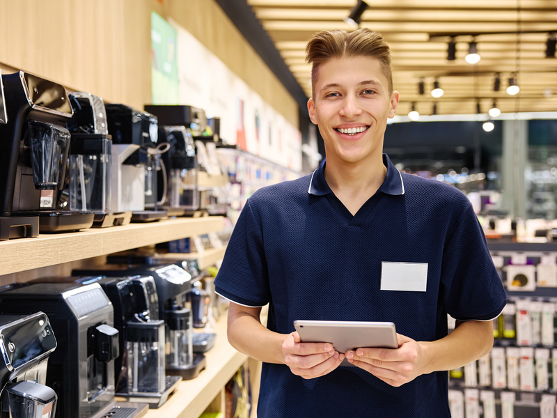 Ein lächelnder Verkäufer steht in einem Geschäft vor einem Regal mit Kaffeemaschinen und hält ein Tablet in den Händen.