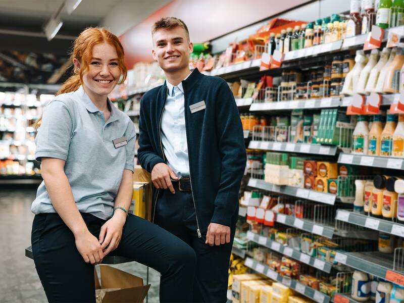 Zwei Personen  stehen lächelnd in einem Supermarkt-Gang mit Regalen voller Produkte im Hintergrund.
