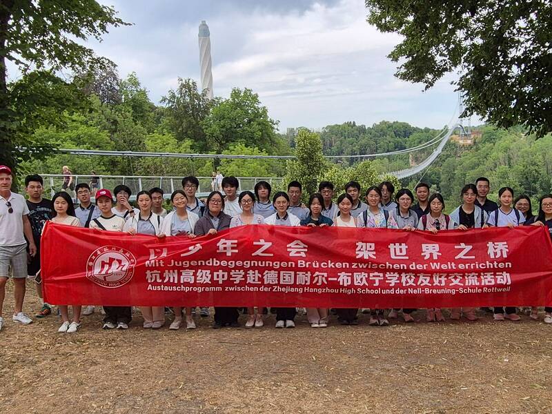 Eine Gruppe von Menschen unterschiedlichen Alters steht im Freien und hält ein langes rotes Banner mit chinesischen und deutschen Schriftzügen, wobei im Hintergrund eine Hängebrücke zu sehen ist.