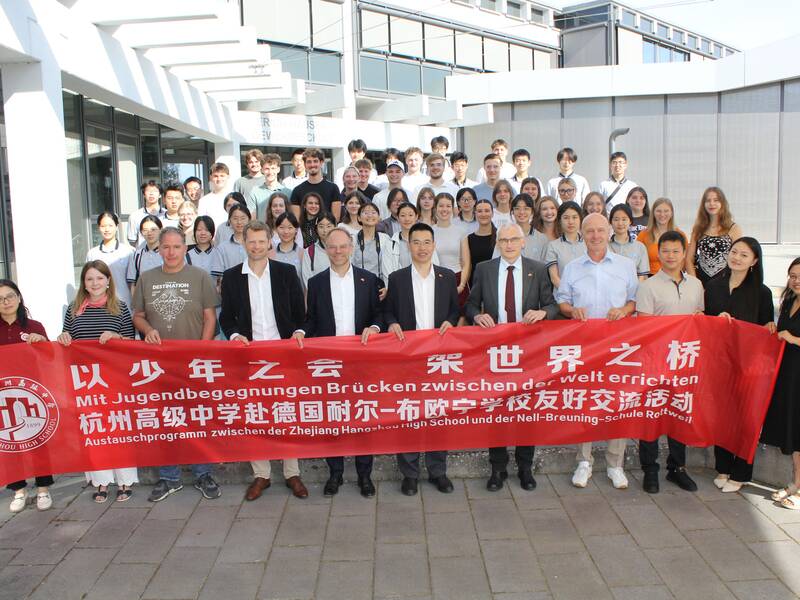 Eine große Gruppe von Personen steht auf einer Treppe vor einem Gebäude und hält ein rotes Banner mit einem deutsch-chinesischen Text, der auf ein Austauschprogramm hinweist.