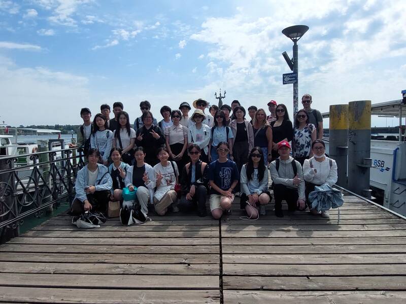 Eine große Gruppe von Menschen posiert gemeinsam für ein Foto auf einem Holzsteg vor einem Gewässer, während im Hintergrund Boote zu sehen sind.