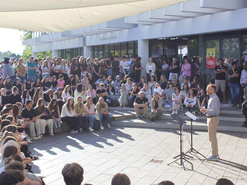 Vor einem Gebäude hält eine Person eine Rede vor einer großen, dicht gedrängten Gruppe von sitzenden und stehenden Zuhörern.
