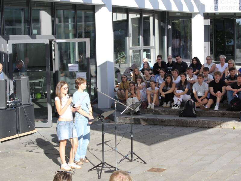 Zwei Personen stehen mit einem Mikrofon im Freien vor einer Gruppe sitzender Menschen, während ein Mann neben Lautsprechern im Hintergrund steht.