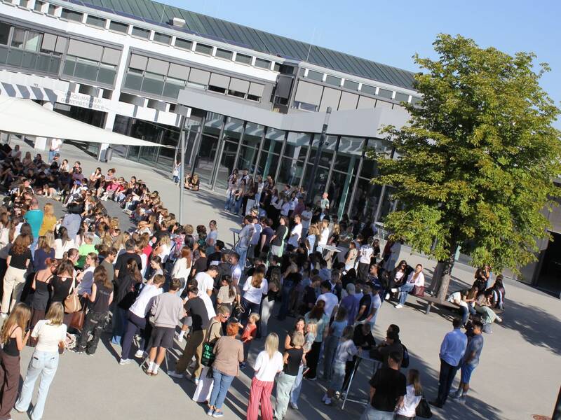 Eine große Gruppe von Menschen versammelt sich auf einem Schulhof vor einem modernen Gebäude mit großen Glasfenstern, während ein Baum Schatten spendet.
