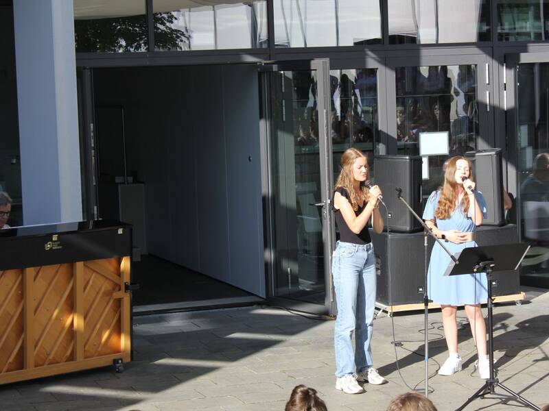 Zwei junge Frauen stehen vor einem Gebäude und singen in Mikrofone, während eine Person am Klavier spielt.