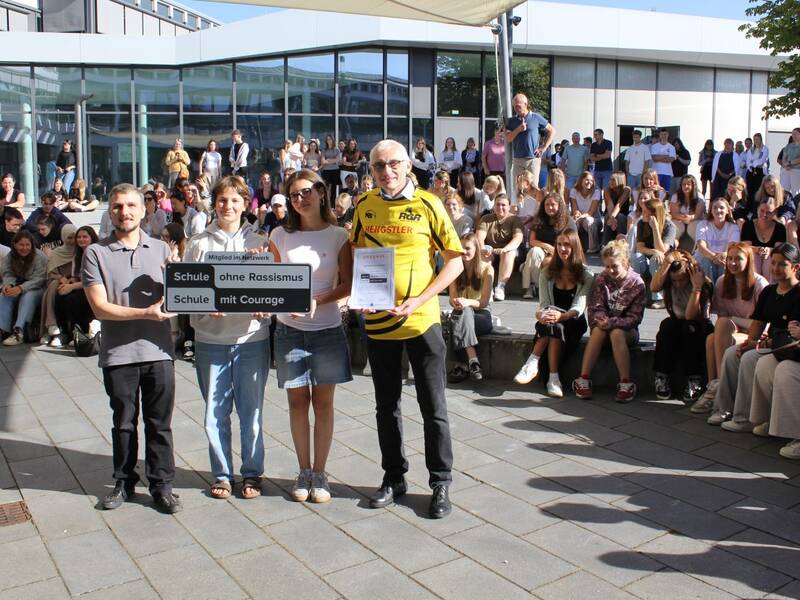 Eine Gruppe von vier Personen hält ein Schild mit der Aufschrift "Schule ohne Rassismus - Schule mit Courage" vor einer größeren Menge von sitzenden Menschen.