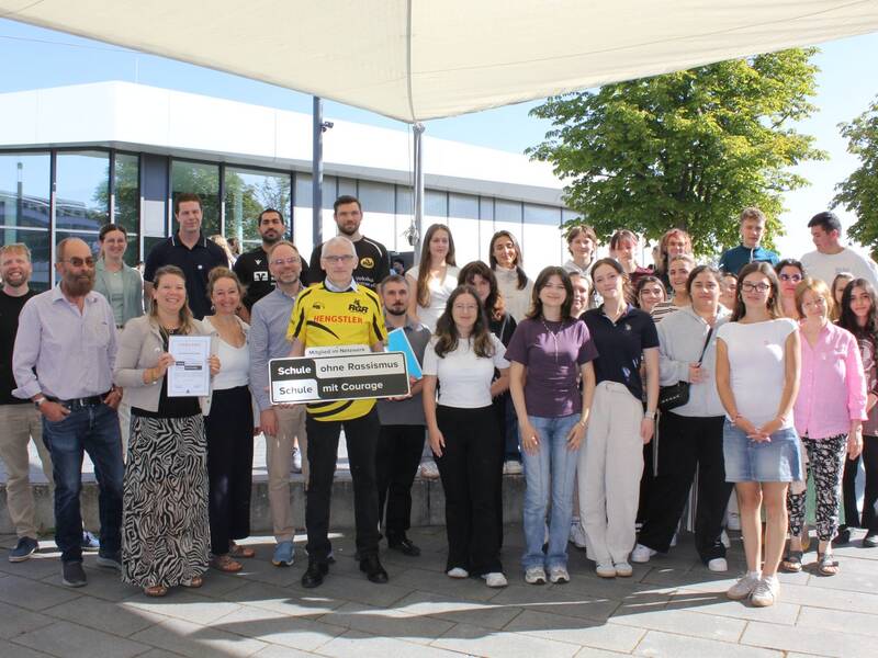 Eine Gruppe von Menschen steht auf einer Treppe im Freien vor einem modernen Gebäude, einige davon halten Schilder mit der Aufschrift "Schule ohne Rassismus - Schule mit Courage".