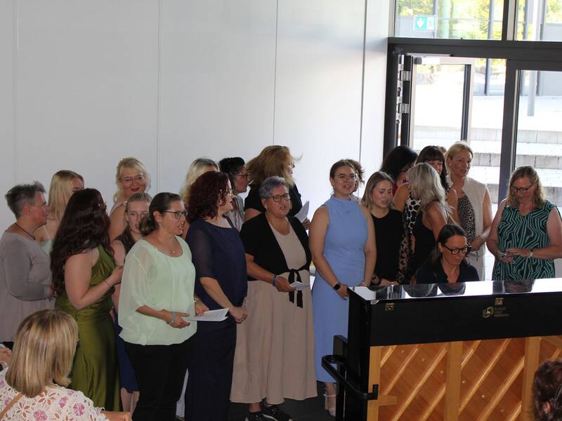 Eine Gruppe von Frauen steht zusammen und singt neben einem Klavier, das von einer Person gespielt wird.