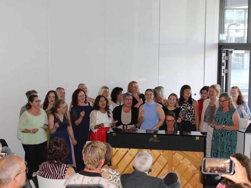 Eine Gruppe von Menschen singt freudig hinter einem Klavier, während jemand im Vordergrund das Geschehen mit einem Handy filmt.