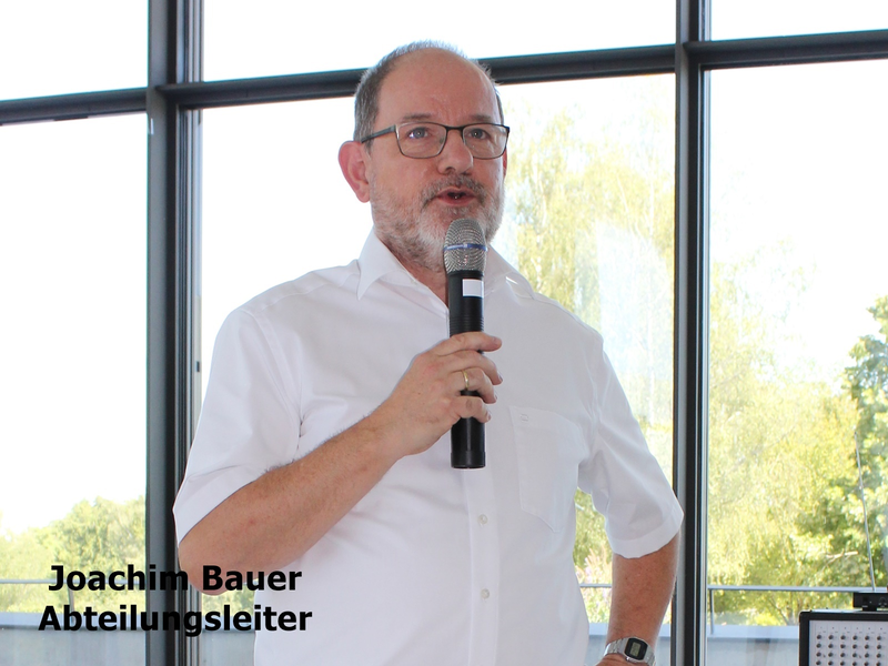 Es ist ein Mann in einem weißen Hemd, der mit einem Mikrofon in der Hand vor großen Fenstern steht.