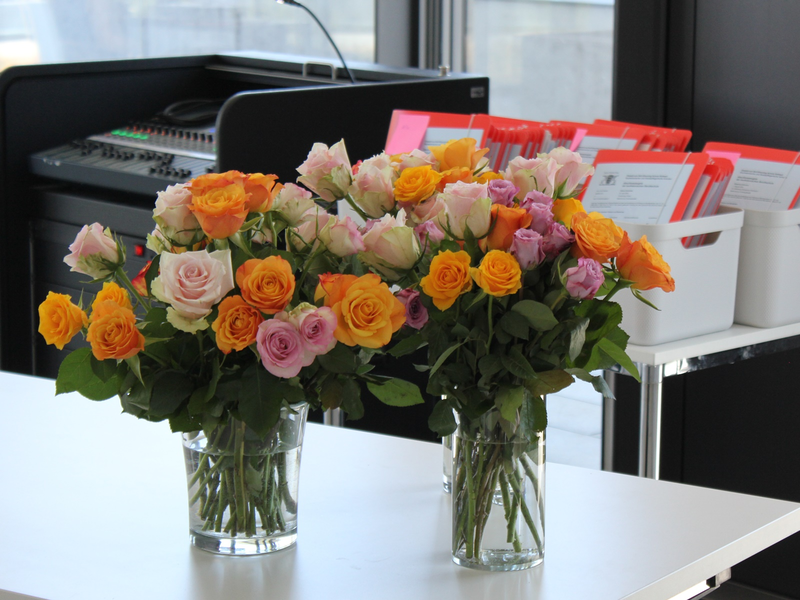 Zwei Vasen mit bunten Rosen stehen auf einem Tisch vor einem Hintergrund mit Technikgeräten und weißen Behältern, die rote Mappen enthalten.
