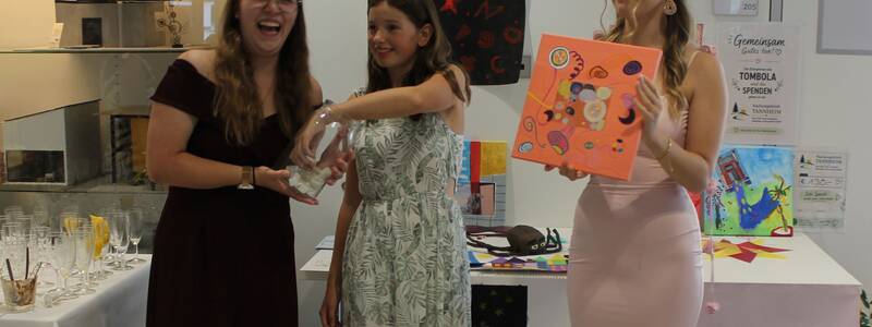 Drei junge Frauen stehen fröhlich zusammen, wobei eine eine Glasschale hält und eine andere ein buntes Kunstwerk präsentiert.