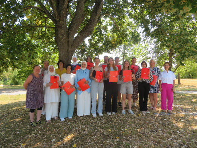 Eine Gruppe von fünfzehn lächelnden Menschen steht im Freien unter einem Baum und hält rote Mappen in den Händen.