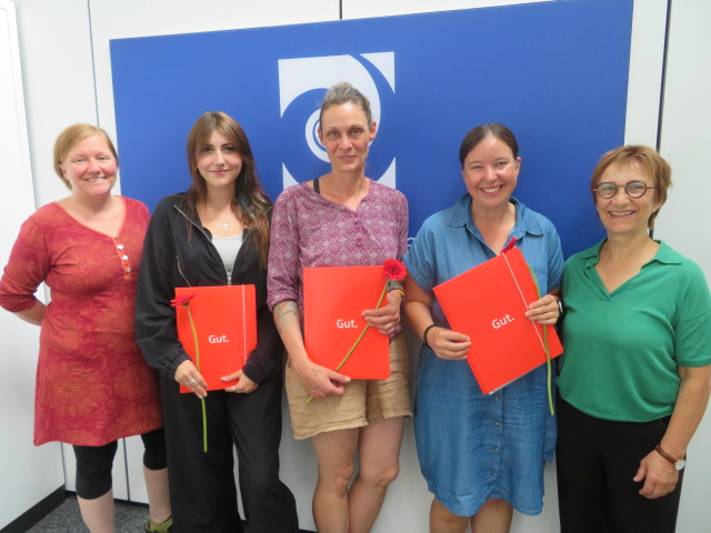 Fünf lächelnde Frauen stehen nebeneinander vor einem blauen Hintergrund, wobei drei von ihnen identische rote Ordner mit der Aufschrift "Gut." und eine Blume in der Hand halten.