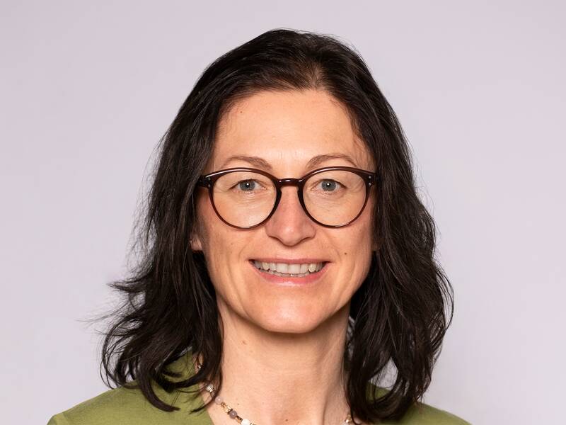 Eine lächelnde Frau mit Brille und dunklem Haar vor einem hellen Hintergrund.