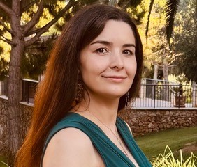 Eine Frau mit langen dunklen Haaren trägt ein ärmelloses, grünes Kleid und steht draußen vor einem Baum.